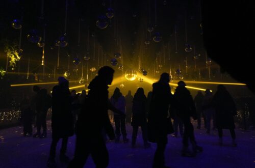 Besišypsantys žmonės vaikščioja šviesų festivalio parke tarp tviskančių girliandų.